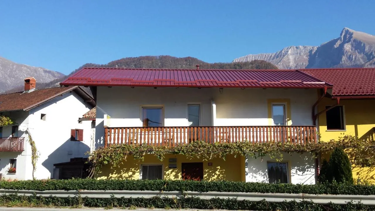 Naša hiša v Idrskem: rdeča streha, lesen balkon z vinsko trto, za njo gore